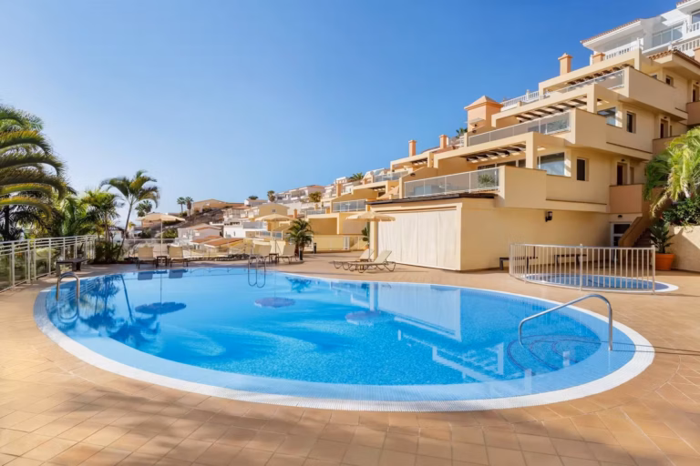 A sunny daytime view of the Wyndham Residences Tenerife Costa Adeje, featuring a large, oval-shaped swimming pool with a light blue rim and crystal-clear water.