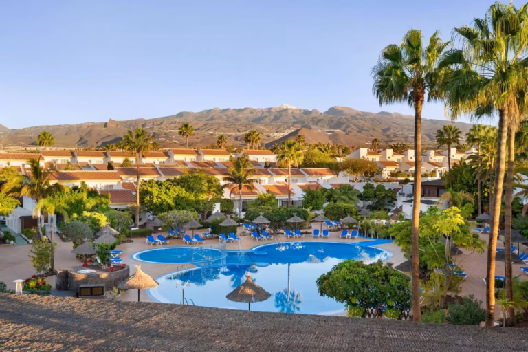An elevated view of the Wyndham Residences Tenerife Golf del Sur, showcasing a large, multi-section swimming pool surrounded by numerous blue lounge chairs and straw umbrellas.